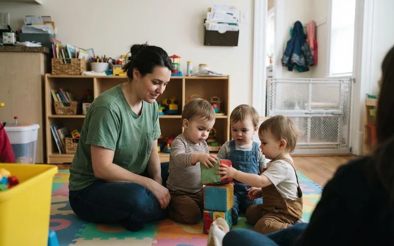 An educator kneeling down to eye level with a small group of toddlers during a supervised indoor play session